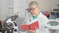 Mature scientist man with grey hair in lab coat studying notes and using microscope in a laboratory setting