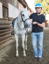 Mature smiling man farmer in helmet standing with white horse at stable Royalty Free Stock Photo