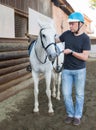 Mature smiling man farmer in helmet standing with white horse at stable Royalty Free Stock Photo