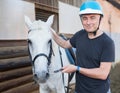 Mature smiling man farmer in helmet standing with white horse at stable Royalty Free Stock Photo