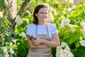 Mature smiling woman in apron with pruners in backyard garden near blooming white hydrangea bush