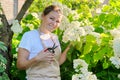 Mature smiling woman in apron with pruners in backyard garden near blooming white hydrangea bush