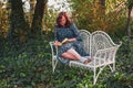 A serious woman of 50 years in retro style is reading a book in a romantic garden with a white decorative antique bench