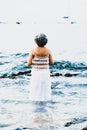 woman with dark gray hair and a white crochet dress standing on rocks in