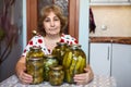 Mature woman embracing glass jars with marinated cucumbers while sitting in own domestic kitchen