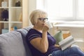 Mature woman in glasses relaxing on couch at home, drinking tea and reading a book alone Royalty Free Stock Photo