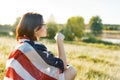 Mature woman listens to music, an audiobook on headphones, relaxes in nature. On the shoulders American flag, background sunset, Royalty Free Stock Photo