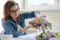 Mature woman making bouquet of lilac branches at home at table