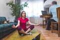 A mature woman performs yoga asana while a man works at a computer at home.