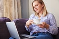 Mature Woman Sitting On Sofa Look At Laptop And Drinking Hot Drink