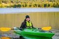 Mature women in green safety life jacket sits in green kayak and look at camera holding yellow paddle across boat.