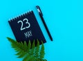 May 23th. Day 23 of month, Calendar date. Black notepad sheet, pen, fern twig, on a blue background