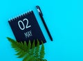 May 2th. Day 2 of month, Calendar date. Black notepad sheet, pen, fern twig, on a blue background.