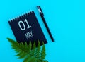 May 1th. Day 1 of month, Calendar date. Black notepad sheet, pen, fern twig, on a blue background.