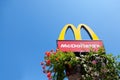 Mc Donald`s logo with its iconic M with a blue sky background and some flowers at its bottom