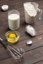 Measuring cup with flour, glass of milk, broken egg and salt, metal whisk on table