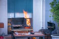 Meat being cooked on barbeque and fire with flames and food and utensils on table