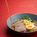 Meat Broth pouring in Ramen soup. Bowl of noodles with meat slices, egg, corn and herbs on red background. Spicy Royalty Free Stock Photo