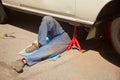 Mechanic in blue dirty uniform lying down and working under car at auto service garage