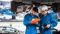 Mechanic in blue workwear uniform checks the vehicle maintenance checklist with his assistant under the lifted car.