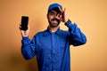 Mechanic man with beard wearing blue uniform and cap holding smartphone showing screen with happy face smiling doing ok sign with Royalty Free Stock Photo
