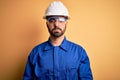 Mechanic man with beard wearing blue uniform and safety glasses over yellow background with serious expression on face