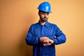 Mechanic man with beard wearing blue uniform and safety helmet over yellow background Checking the time on wrist watch, relaxed