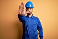 Mechanic man with beard wearing blue uniform and safety helmet over yellow background doing stop sing with palm of the hand