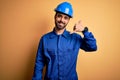 Mechanic man with beard wearing blue uniform and safety helmet over yellow background smiling doing phone gesture with hand and Royalty Free Stock Photo