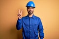 Mechanic man with beard wearing blue uniform and safety helmet over yellow background smiling looking to the camera showing Royalty Free Stock Photo