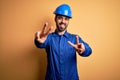 Mechanic man with beard wearing blue uniform and safety helmet over yellow background smiling with tongue out showing fingers of Royalty Free Stock Photo