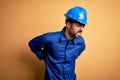 Mechanic man with beard wearing blue uniform and safety helmet over yellow background Suffering of backache, touching back with