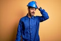 Mechanic man with beard wearing blue uniform and safety helmet over yellow background surprised with hand on head for mistake,