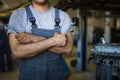 A mechanic in overalls with tools in his hands stands with his arms crossed on his chest.