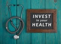 Blackboard with text & x22;Invest in your health& x22; and stethoscope on blue wooden background