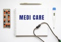 MEDI CARE is written in a notebook on a white table next to pills and a stethoscope