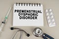 On a gray surface there is a syringe, a stethoscope and a notepad with the inscription - premenstrual dysphoric disorder