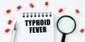 On a white surface are tablets, a pen, a magnifying glass and a notepad with the inscription - TYPHOID FEVER