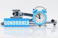 On a white surface there is an alarm clock, a stethoscope and a blue block with the inscription - GONORRHEA