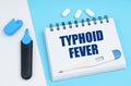 On a white-blue surface are pills, a thermometer, a marker and a notebook with the inscription - TYPHOID FEVER