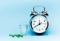 Medical green capsule pills, glass water and alarm clock on blue background. Royalty Free Stock Photo