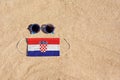 A medical mask in the color of the Croatia flag lies on the sandy beach next to the glasses.