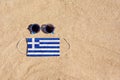 A medical mask in the color of the Greece flag lies on the sandy beach next to the glasses.