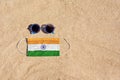 A medical mask in the color of the India flag lies on the sandy beach next to the glasses.