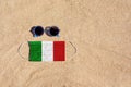 A medical mask in the color of the Italian flag lies on the sandy beach next to the glasses.