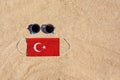 A medical mask in the color of the Turkish flag lies on the sandy beach next to the glasses.