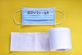 Medical mask with inscription covid-19 and roll of unwound toilet paper on yellow background. Body protection, hygiene products