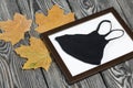 Medical mask in a photo frame. Among the dried maple leaves. On brushed pine boards painted black and white Royalty Free Stock Photo