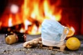 Medical mask on a white cup of tea with lemon, ginger, orange and jam against the background of a burning fireplace. Healthcare.