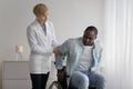 Serious female therapist in white coat looks at mature african american to disabled man